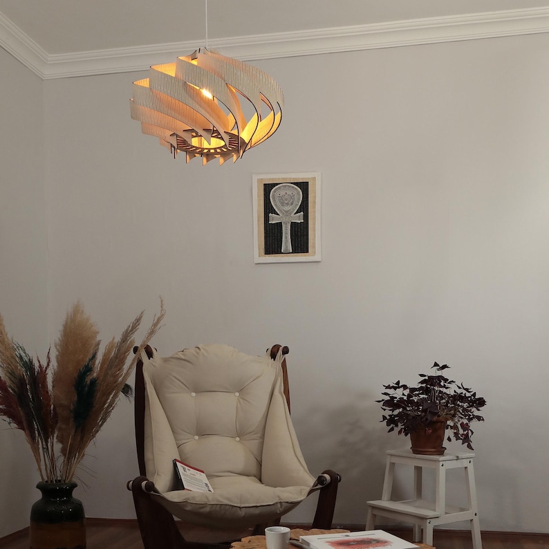 May include: A decorative wooden pendant light with a layered design hangs from the ceiling. The light fixture is made of light-coloured wood and emits a warm glow. The room also features a beige armchair, a framed artwork, and a potted plant.