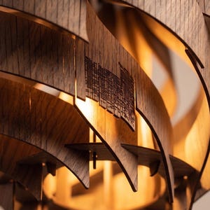 May include: Close-up of a modern wooden pendant light fitting. The fitting features layered, curved wooden slats in a warm brown tone, with a cityscape design cut into some of the slats. The light emits a warm glow.