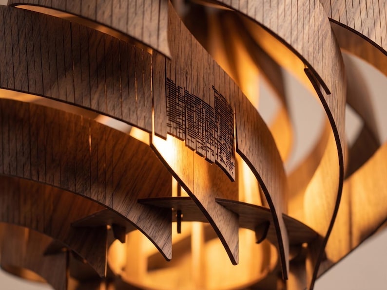 May include: Close-up of a modern wooden pendant light fitting. The fitting features layered, curved wooden slats in a warm brown tone, with a cityscape design cut into some of the slats. The light emits a warm glow.