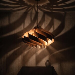 May include: A wooden pendant light with a layered, curved design. The light casts a patterned shadow on the wall. A small vase sits on a dark surface below the light.