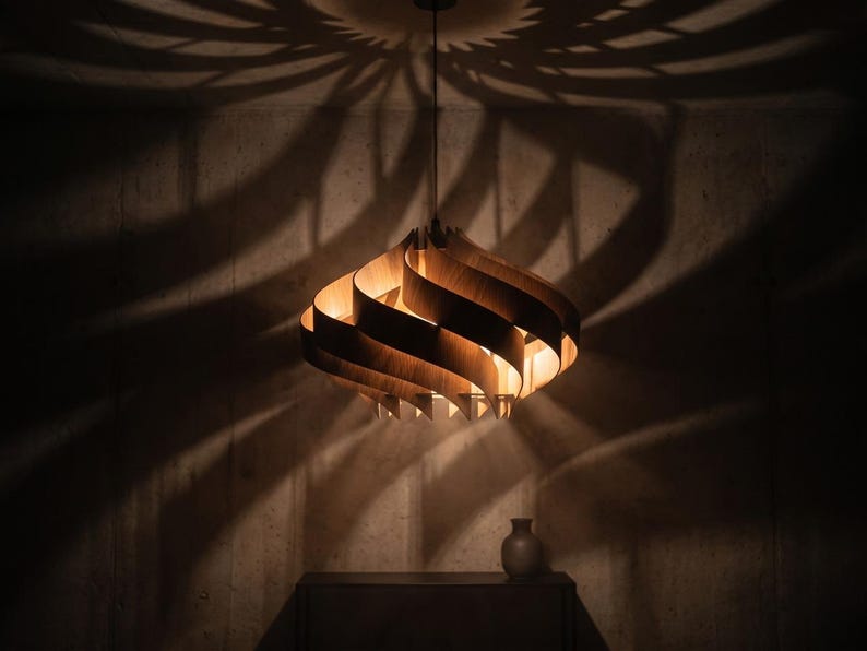May include: A wooden pendant light with a layered, curved design. The light casts a patterned shadow on the wall. A small vase sits on a dark surface below the light.