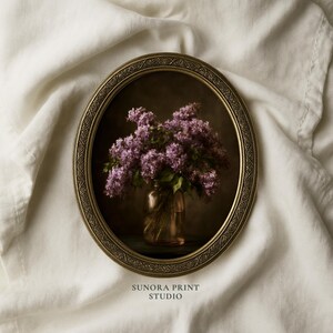 Peut inclure: Un cadre ovale orné de couleur dorée contenant une impression de nature morte représentant un vase en verre rempli de fleurs de lilas violettes. Le texte SUNORA PRINT STUDIO est imprimé en petites lettres sombres sous le cadre.