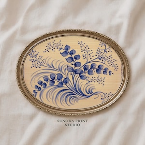 May include: An oval-shaped wall art piece featuring a beige background with a blue botanical illustration of leaves and sprigs. The artwork is encased in a metallic gold frame with a textured, beaded edge. The text SUNORA PRINT STUDIO is visible.