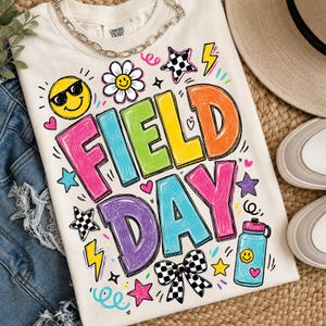 Può includere: T-shirt color crema con la scritta "FIELD DAY" in lettere colorate e bordate. Il design include una faccina sorridente, stelle, fulmini e una borraccia. Una collana a catena argentata è drappeggiata sulla maglietta.