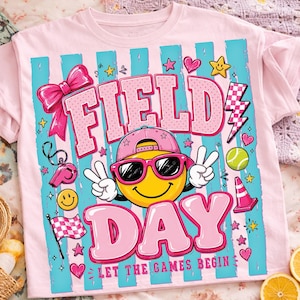 Può includere: T-shirt rosa chiaro con un design grafico colorato. Il design presenta le parole "FIELD DAY" in lettere rosa, una faccina gialla con occhiali da sole e berretto, stelle, cuori e un fulmine. Il testo "LET THE GAMES BEGIN" è in basso.