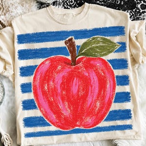 May include: A cream-colored t-shirt featuring a large, textured red apple graphic with a brown stem and a green leaf. The apple is centered over a background of horizontal, distressed blue brushstroke stripes that span the width of the shirt.