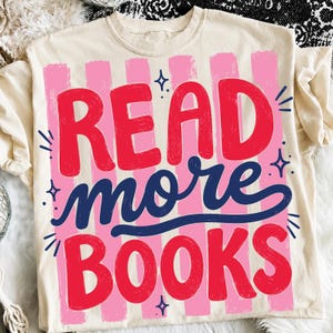 May include: A cream-colored t-shirt featuring a graphic design with vertical pink stripes. The text READ and BOOKS is printed in a bold red font, while the word more is written in a dark blue script. Small dark blue star icons surround the text.