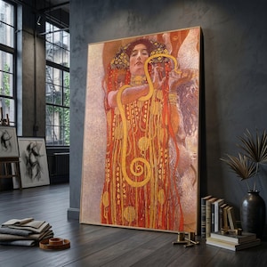 May include: A framed art print of a Gustav Klimt painting. The artwork features a woman in a red and gold dress with a golden snake. The painting has a detailed, ornate style with a warm color palette. The print is displayed in a room with dark walls and hardwood floors.