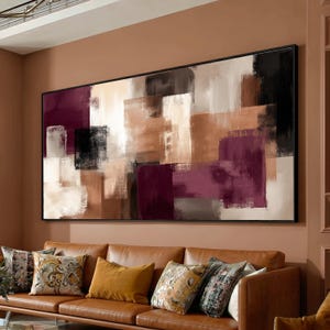 May include: Abstract art piece with a black frame. The artwork features a mix of rectangular shapes in shades of burgundy, brown, beige, and black. The painting hangs above a brown leather sofa with decorative pillows.