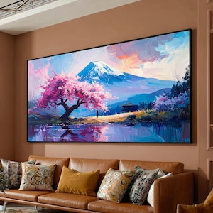 Può includere: Un grande dipinto incorniciato raffigura un sereno paesaggio giapponese. Una montagna innevata si erge sopra un lago, con un vibrante albero di fiori di ciliegio rosa in primo piano. L'opera d'arte presenta un piccolo edificio e una figura, con un cielo colorato.