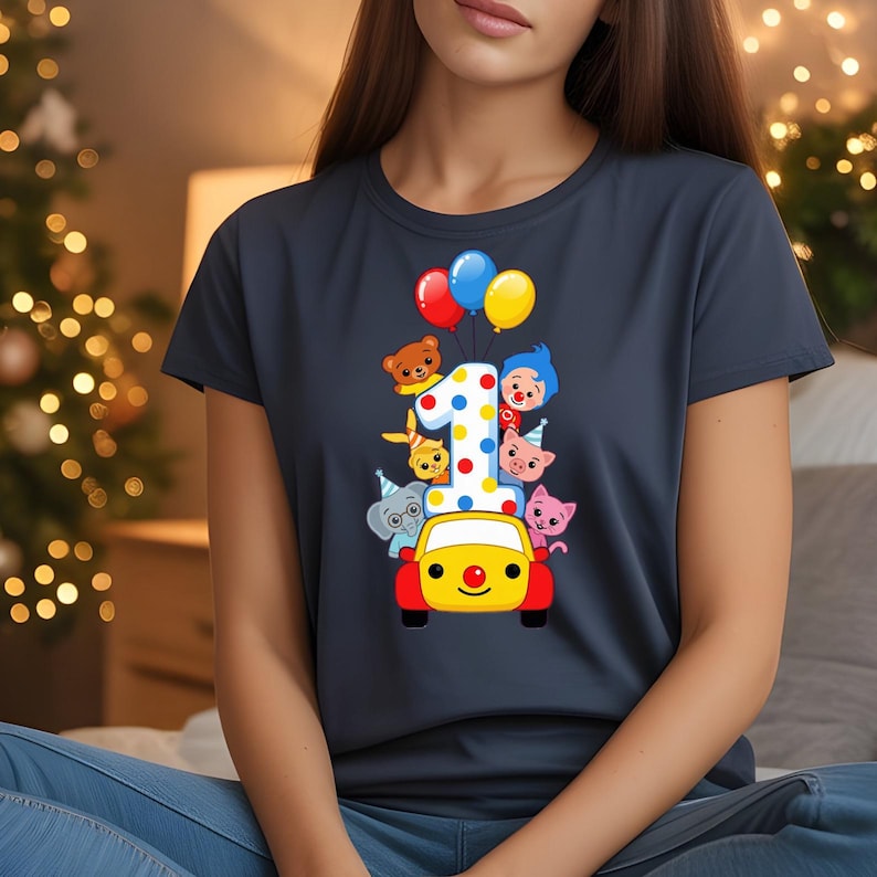K&ouml;nnte beinhalten: Marineblaues T-Shirt mit einer farbenfrohen Illustration f&uuml;r den ersten Geburtstag. Das Design umfasst die Zahl 1 mit Punkten, Zeichentricktieren, Luftballons und ein gelbes Auto mit roter Nase und R&auml;dern. Das Shirt ist ein l&auml;ssiger Stil.
