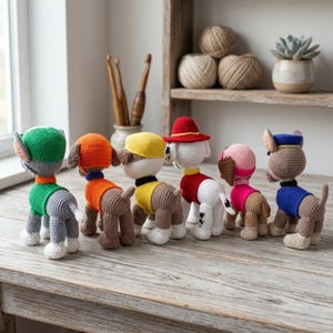 K&ouml;nnte beinhalten: Ein Set aus sechs geh&auml;kelten Hundefiguren in verschiedenen Farben, darunter Gr&uuml;n, Orange, Gelb, Rot, Pink und Blau. Jede Figur tr&auml;gt einen eigenen Hut und eine Weste, steht auf einer Holzoberfl&auml;che und zeigt detaillierte Garnstrukturen.
