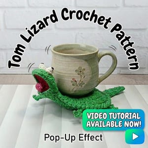Könnte beinhalten: Ein grüner gehäkelter Eidechsen-Untersetzer, der eine beige Keramiktasse mit Blumendesigns trägt. Der Text lautet "Tom Lizard Crochet Pattern" und "Video Tutorial Available Now!" Der Untersetzer hat einen Pop-up-Effekt.
