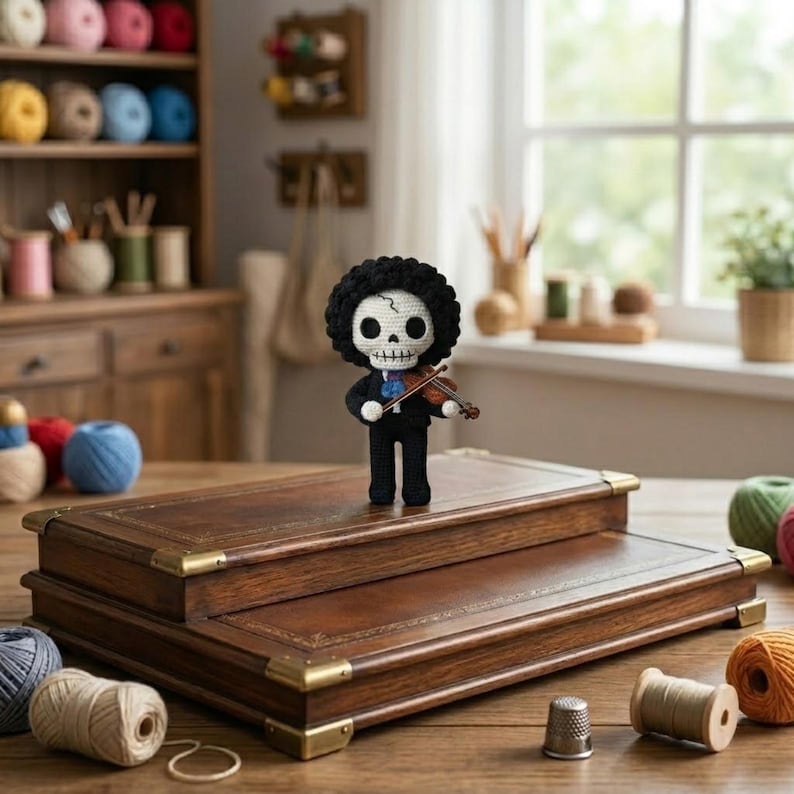 Pode incluir: Figura de croch&ecirc; de um esqueleto com afro preto, vestindo um terno preto e tocando violino. A figura est&aacute; em uma caixa de couro marrom de dois n&iacute;veis com cantos de lat&atilde;o. Fios e acess&oacute;rios de costura espalhados.