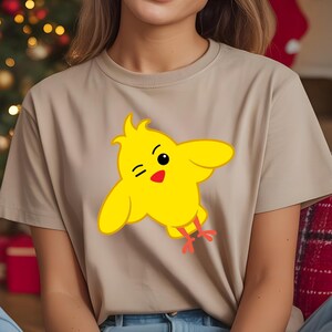 K&ouml;nnte beinhalten: Beiges T-Shirt mit einem gelben Cartoon-K&uuml;ken-Design. Das K&uuml;ken hat schwarze Augen, einen roten Schnabel und rote F&uuml;&szlig;e. Es zwinkert mit ausgebreiteten Fl&uuml;geln.