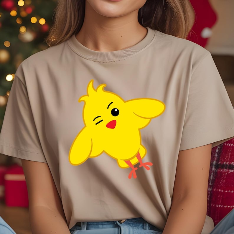 K&ouml;nnte beinhalten: Beiges T-Shirt mit einem gelben Cartoon-K&uuml;ken-Design. Das K&uuml;ken hat schwarze Augen, einen roten Schnabel und rote F&uuml;&szlig;e. Es zwinkert mit ausgebreiteten Fl&uuml;geln.