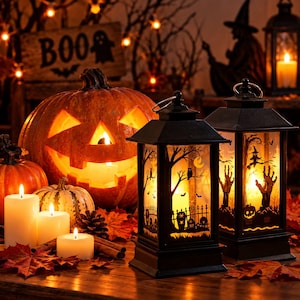 May include: Two black rectangular lanterns with illuminated Halloween scenes featuring silhouettes of trees, owls, tombstones, and a witch. The lanterns are displayed alongside a carved jack-o'-lantern, smaller pumpkins, white pillar candles, and autumn leaves.