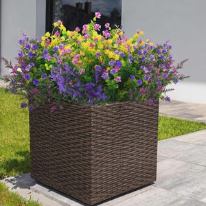 Pode incluir: Uma floreira quadrada de vime castanho cheia de flores artificiais coloridas. As flores s&atilde;o uma mistura de amarelo, rosa e roxo, criando uma exibi&ccedil;&atilde;o vibrante. A floreira est&aacute; sobre uma superf&iacute;cie de pedra cinzenta.