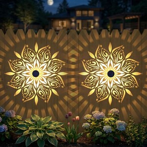 May include: Two solar-powered decorative lights with a floral design are mounted on a wooden fence. The lights cast intricate patterns on the fence. The scene is set at night, with a full moon and a house in the background, and flowers in the foreground.