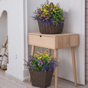 Pode incluir: Duas cestas de vime castanhas cheias de flores artificiais coloridas. Uma cesta est&aacute; numa mesa lateral de madeira clara com uma gaveta, e a outra est&aacute; no ch&atilde;o. As flores incluem amarelo, roxo, rosa e verde.