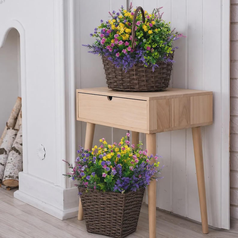 Pode incluir: Duas cestas de vime castanhas cheias de flores artificiais coloridas. Uma cesta est&aacute; numa mesa lateral de madeira clara com uma gaveta, e a outra est&aacute; no ch&atilde;o. As flores incluem amarelo, roxo, rosa e verde.