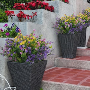 Pode incluir: Tr&ecirc;s floreiras tecidas cinza escuro cheias de flores artificiais coloridas em tons de roxo, amarelo e rosa. As floreiras est&atilde;o em uma escada de tijolos vermelhos ao lado de uma parede de concreto. Outras plantas s&atilde;o vis&iacute;veis ao fundo.