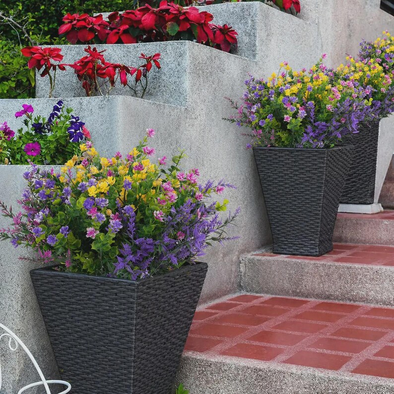 Pode incluir: Tr&ecirc;s floreiras tecidas cinza escuro cheias de flores artificiais coloridas em tons de roxo, amarelo e rosa. As floreiras est&atilde;o em uma escada de tijolos vermelhos ao lado de uma parede de concreto. Outras plantas s&atilde;o vis&iacute;veis ao fundo.