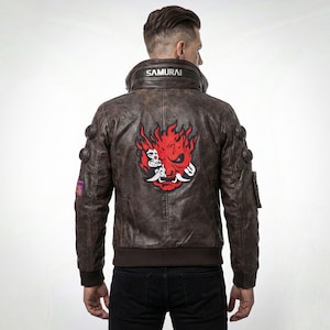 Puede incluir: Chaqueta de cuero marrón con acabado desgastado, con un gran gráfico rojo y blanco en la espalda. La chaqueta tiene un dobladillo acanalado, múltiples bolsillos y la palabra "SAMURAI" en el cuello.