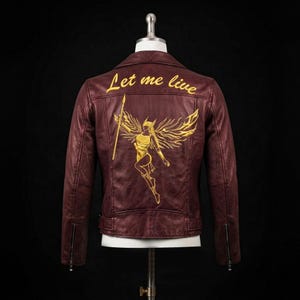 May include: A dark maroon leather jacket featuring gold embroidery on the back. The design includes a winged figure holding a spear and the text Let me live in a cursive script. The jacket has long sleeves with zippered cuffs and a collar.