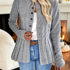 May include: A grey knit cardigan featuring a cable knit pattern on the front panels, a button-down closure with dark buttons, and a flared, pleated hemline. The garment is worn over a white top and blue denim jeans with a brown leather belt.