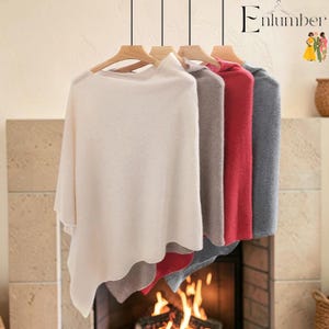 May include: A collection of four ponchos hanging on wooden hangers, displayed in a row. The ponchos are available in solid cream, light gray, red, and dark gray colors, each featuring a soft, draped silhouette and a simple, clean design.