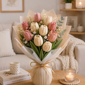 May include: A bouquet of crocheted tulips in shades of pink and cream, accented with green knitted leaves and small white filler flowers. The flowers are wrapped in translucent paper and tied with a raffia bow, displayed in a ribbed cream ceramic vase.