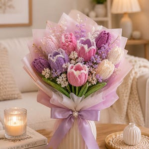 May include: A bouquet of crocheted tulips in shades of pink, purple, and white, accented with small white and lavender filler flowers and green stems. The flowers are wrapped in translucent pink paper and tied with a lavender ribbon in a white vase.