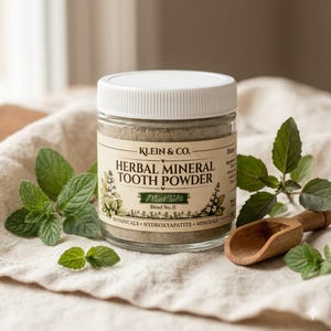 May include: A clear glass jar with a white lid containing grey herbal mineral tooth powder. The label reads Klein & Co. Herbal Mineral Tooth Powder Mint Tulsi Blend No. II. The jar is surrounded by fresh green mint leaves and a small wooden scoop.