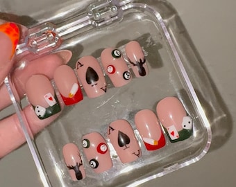 May include: A set of ten press-on nails in a clear plastic storage case. The nails feature a nude base with various designs including black spade symbols, billiard balls, red and white playing cards, and black silhouettes of deer heads.