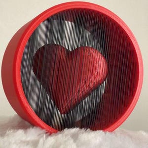 Puede incluir: Escultura roja en forma de corazón con efecto 3D, dentro de un marco circular rojo con líneas negras verticales. La escultura está sobre una superficie blanca y esponjosa. Objeto decorativo.