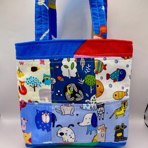 May include: A patchwork tote bag with blue handles and a blue top border. The bag features various fabric panels with animal and nature-themed patterns, including cats, dogs, fish, and trees. The bag is designed for carrying items.