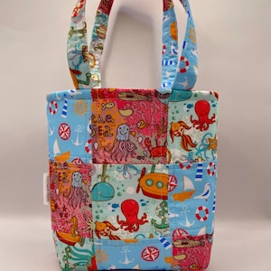 Peut inclure: Un sac fourre-tout en tissu matelassé avec un motif patchwork représentant des éléments marins comme des pieuvres, des baleines, des ancres et des voiliers dans des tons vifs de bleu, rose et jaune. Une étiquette blanche indique Patchwork Panda.