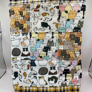 May include: A fabric tote bag with a patchwork design showcasing numerous cat illustrations in various colours. The top and bottom edges are trimmed with a yellow and navy blue gingham pattern. The bag is rectangular in shape.
