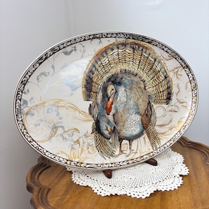 Puede incluir: Una bandeja de servicio ovalada de cerámica con una ilustración central de un pavo con las plumas de la cola desplegadas. La bandeja tiene un fondo color crema con detalles florales dorados y grises y un borde decorativo marrón oscuro.
