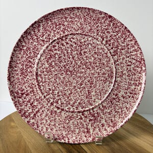 May include: A round dinner plate featuring a speckled pattern of deep maroon or burgundy paint splatters against a creamy white background. The plate has a distinct circular inner rim that separates the center from the wider outer border.