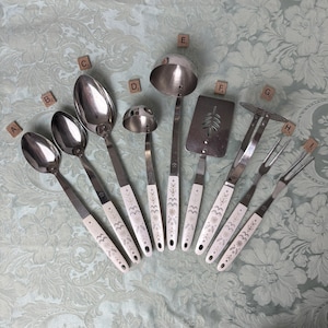May include: A set of nine stainless steel kitchen utensils with white handles featuring a dark floral and geometric pattern. The collection includes various spoons, a ladle, a slotted turner, a masher, and a two-pronged fork, each labeled A through I.
