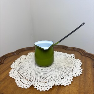 May include: A small olive green enamel pot with a white interior and a long, thin black handle. The pot features a rounded body and a small pouring spout on the rim, designed for heating liquids or melting small quantities of ingredients.