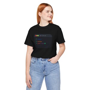 Può includere: T-shirt nero con una grafica di una finestra del terminale del codice. Il testo nella finestra recita "commit", "coffee" e "push code". La parte superiore della finestra dice "dev-life.sh".