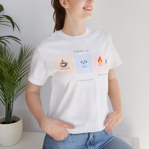 May include: A light grey t-shirt featuring the text "POWERED BY" above three coloured squares. The squares display icons of a coffee cup, code brackets, and a flame, with the words "CAFFEINE", "CODE", and "CHAOS" below. The text "// a developer's formula" is below.