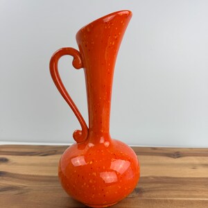 May include: A bright orange ceramic pitcher with a speckled finish, featuring a bulbous base, a tall slender neck, and a curved handle that attaches to the side of the neck and the top of the rounded body, creating a distinct silhouette.