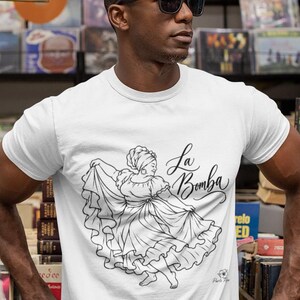 May include: A white short-sleeved t-shirt featuring a black line art graphic of a person in a ruffled dress dancing. The text "La Bomba" is printed in a cursive script above the illustration. The shirt has a crew neckline and a standard fit.