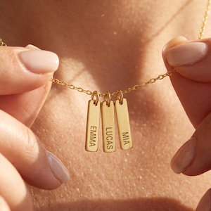 May include: A gold-toned necklace featuring three vertical rectangular bar pendants suspended from a delicate chain. Each bar is engraved with a name in capital letters: EMMA, LUCAS, and MIA, arranged side by side on the chain.