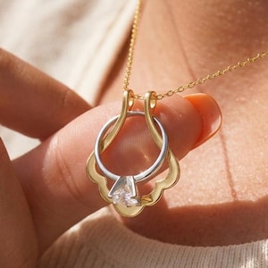 May include: A gold-coloured necklace featuring a pendant designed as a ring. The ring pendant has a silver band and a clear, faceted stone. The pendant hangs from a fine gold chain. The necklace is being held by a person.