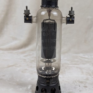 Puede incluir: Una válvula de vacío Telefunken vintage con un cuerpo de vidrio transparente y una base de metal negro. El tubo tiene el texto "Telefunken D.R.P. Typ RS 282" impreso. Dos pequeños conectores metálicos están en los lados, etiquetados "K" y "G".